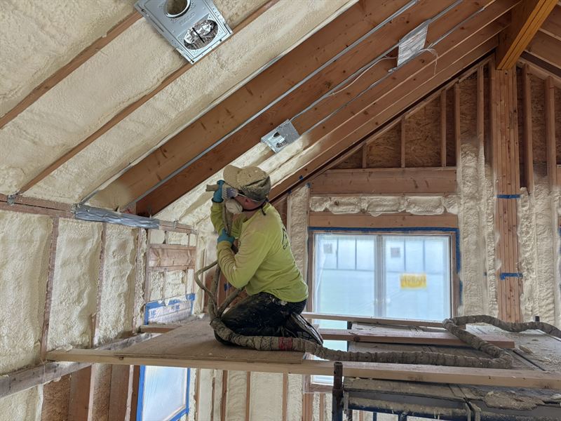 Installer in hi-vis and respirator spraying an attic rafter bay.