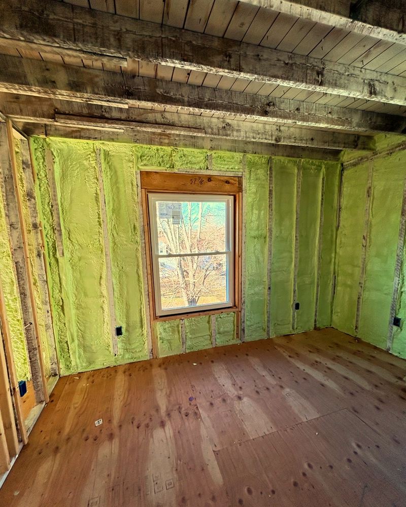 Room in a gut remodel with walls fully insulated in foam around an existing window and hardwood floor.