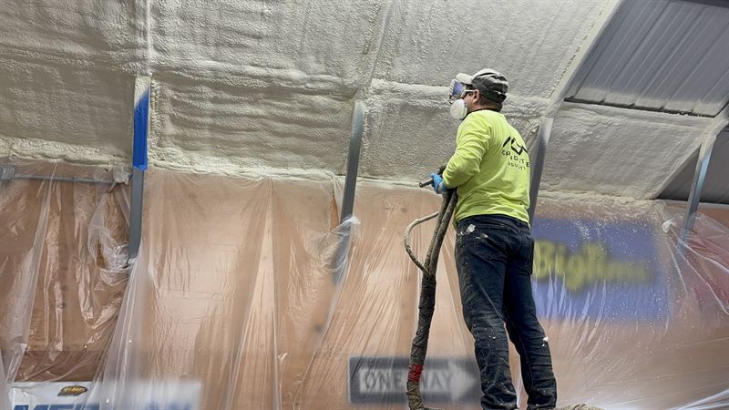 Installer spraying foam along the ceiling line of a masked metal building.